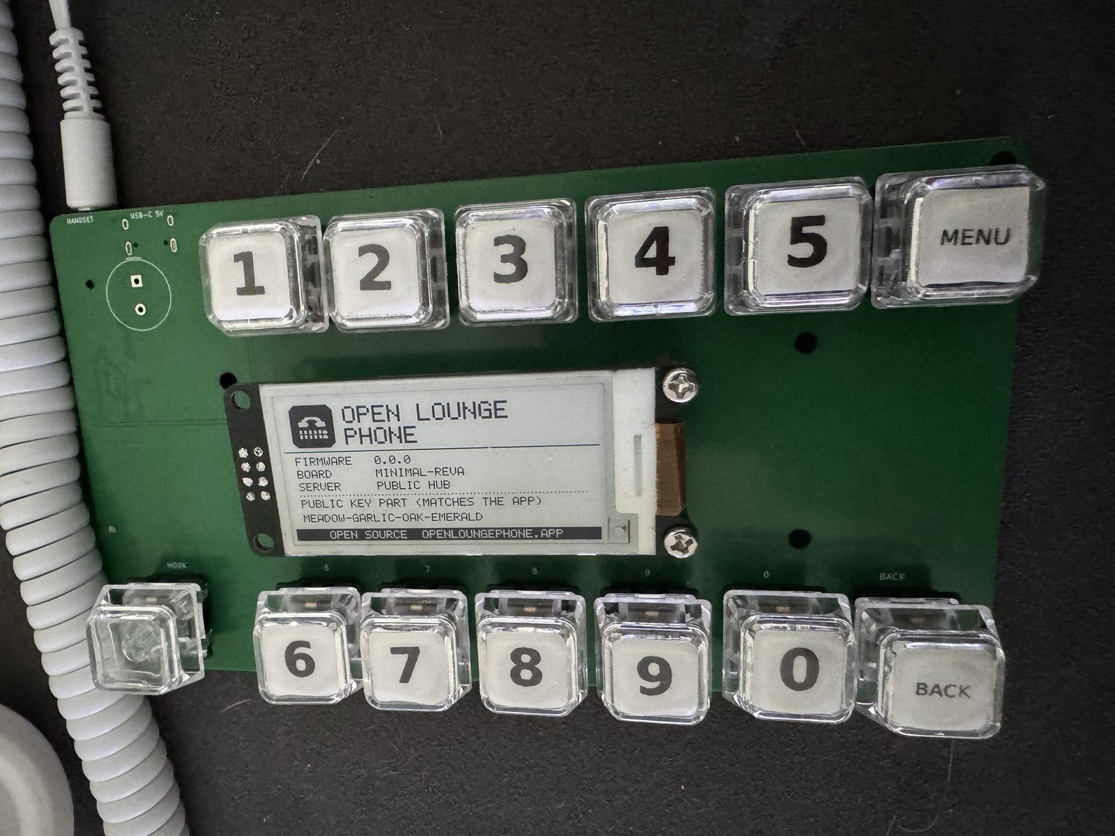 Close-up of the dev board's e-paper display showing Open Lounge Phone, firmware 0.0.0, board minimal-revA, server public hub, and the four words of its key
