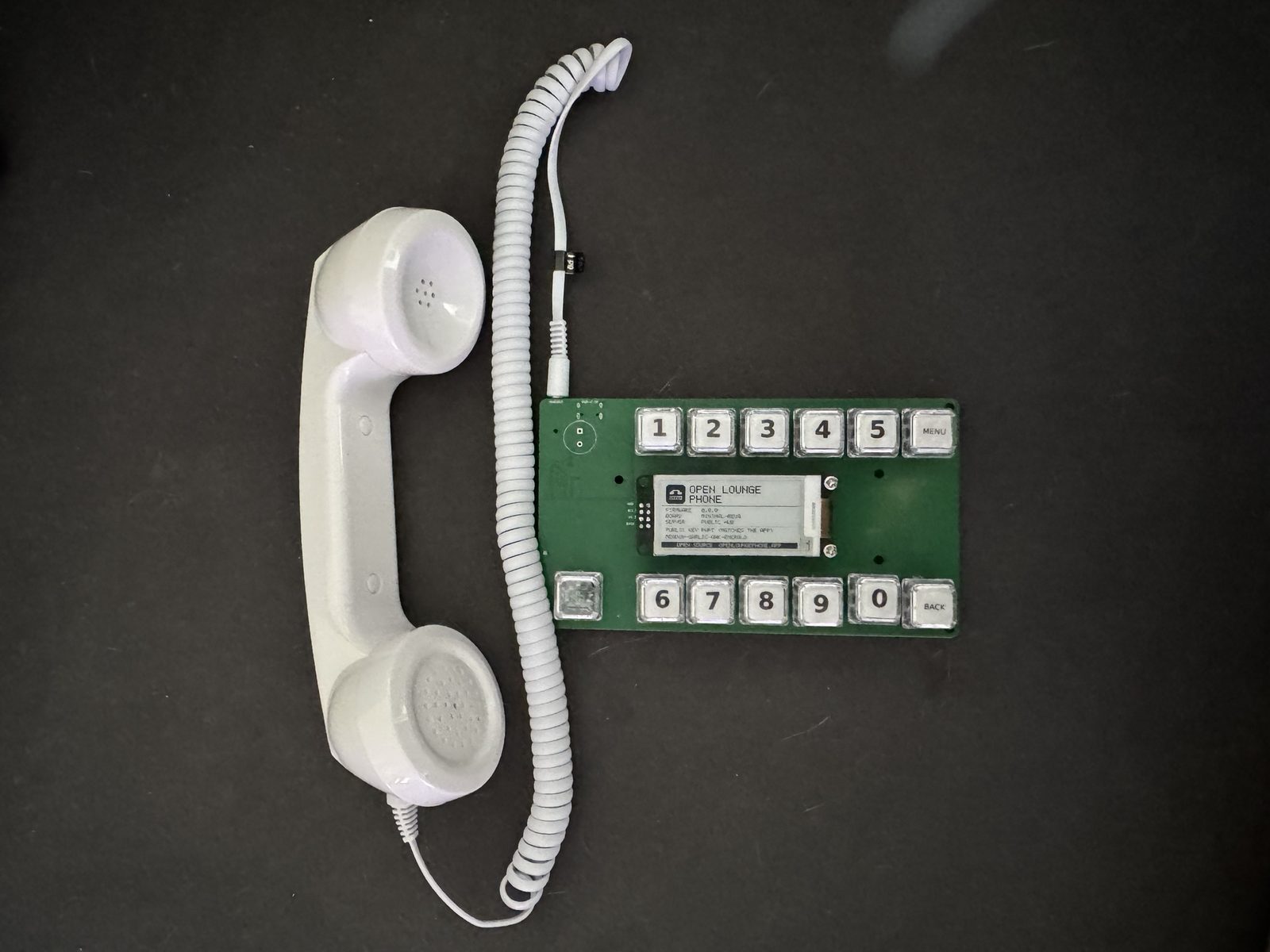A green v0.1 dev board with twelve clear keycaps in two rows, an e-paper display between them, and a white handset on a coiled cord plugged into its jack
